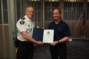 A police officer in a white shirt stands beside Andy Bates with a certificate of appreciation indoors
