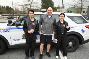 Two youth academy students and a police officer in casual clothing smile beside a police SUV outdoors